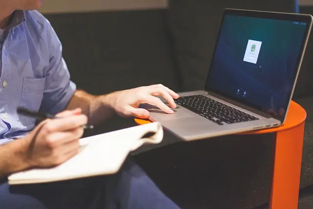 Person am Laptop mit Heft auf den Beinen