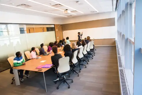 Frau steht vor einem Whiteboard und andere Personen sitzen um einen großen Tisch herum.