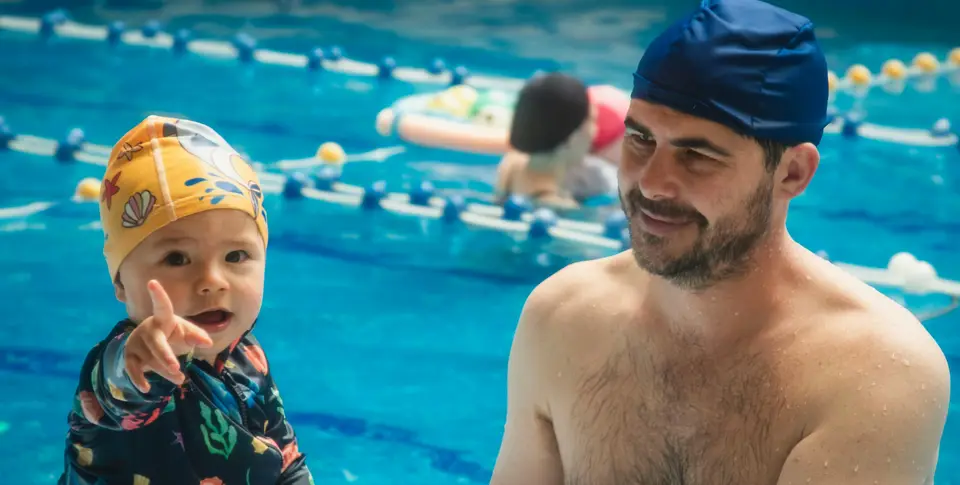 Ein Mann ist mit einem kleinen Kind im Schwimmbad. Das Kind trägt eine gelbe Badekappe und zeigt fröhlich nach vorne.