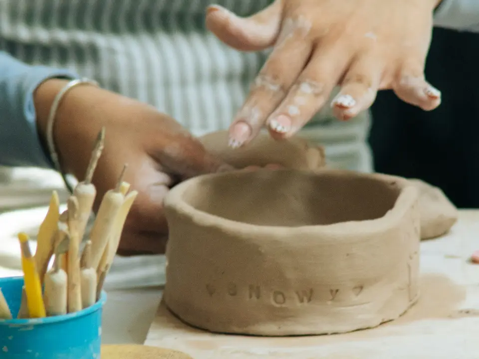 Hands shaping a clay bowl at a workshop table with sculpting tools.