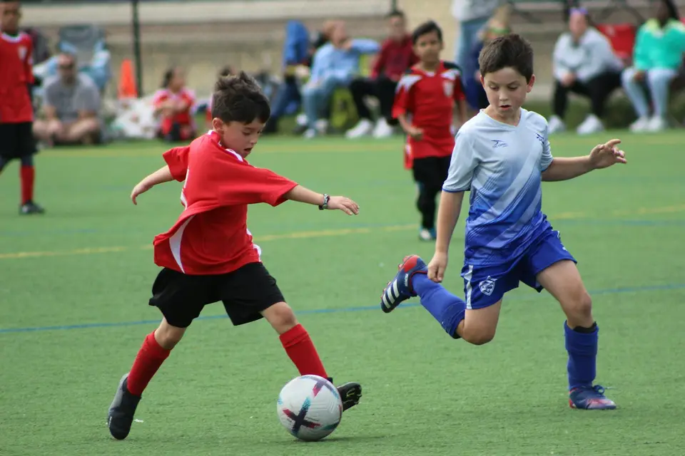 Two young players running after the ball while families watch.