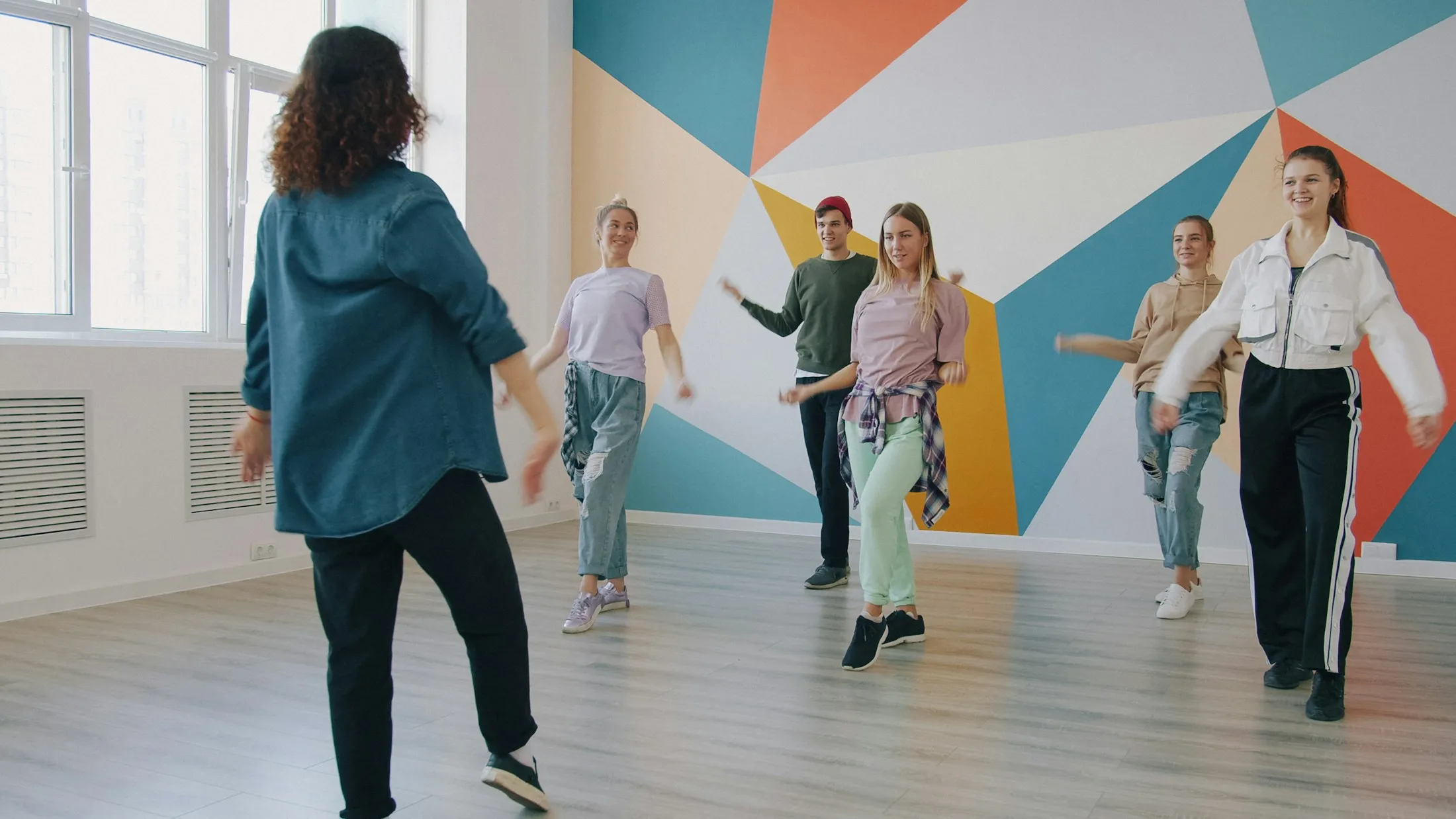 Eine Tanzlehrerin leitet eine Gruppe vor einer bunten Wand an.