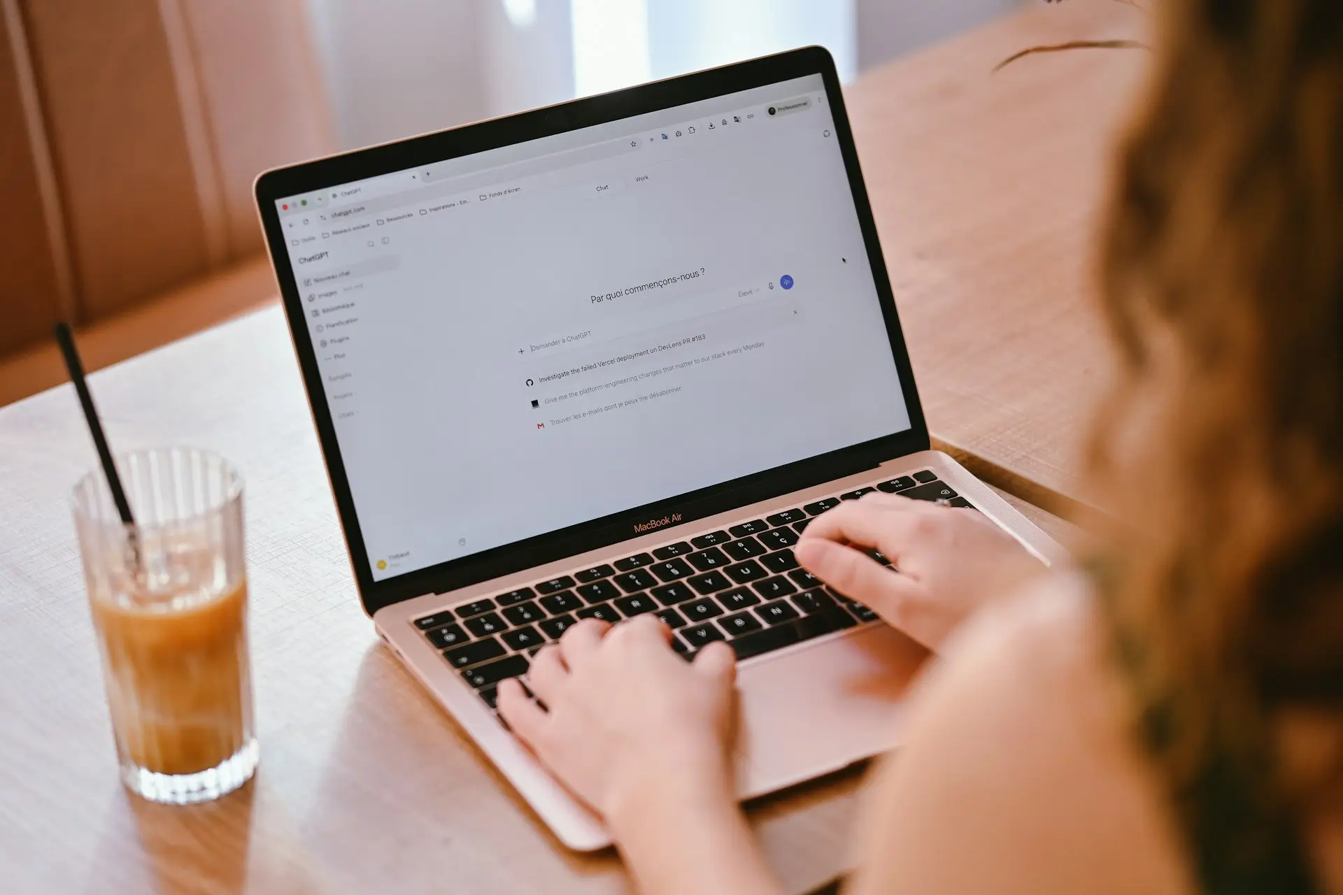 Eine Person tippt an einem Laptop eine Anfrage in einen KI-Chat, daneben steht ein Glas Eiskaffee