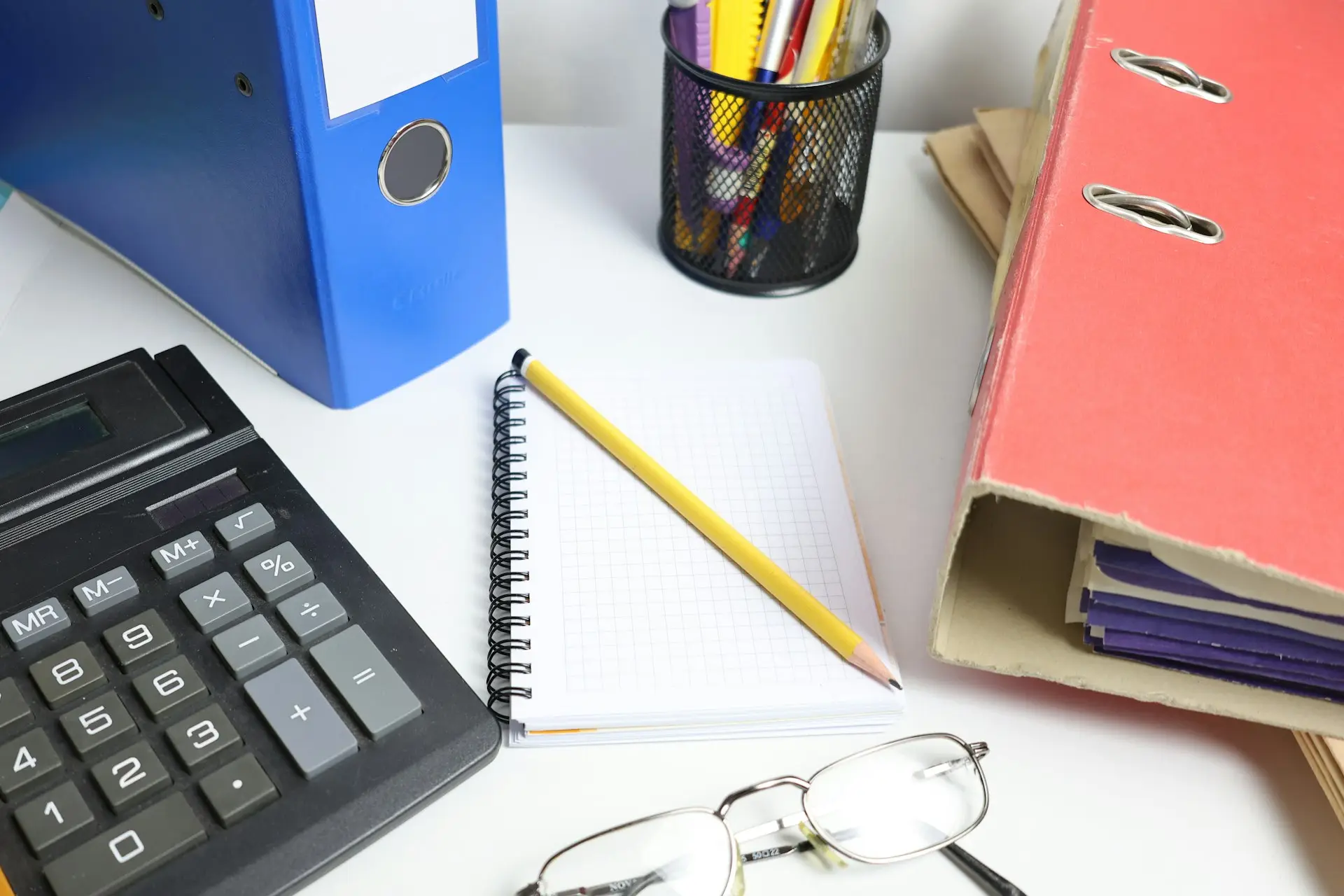 Schreibtisch mit Aktenordnern, Taschenrechner, Notizblock und Brille
