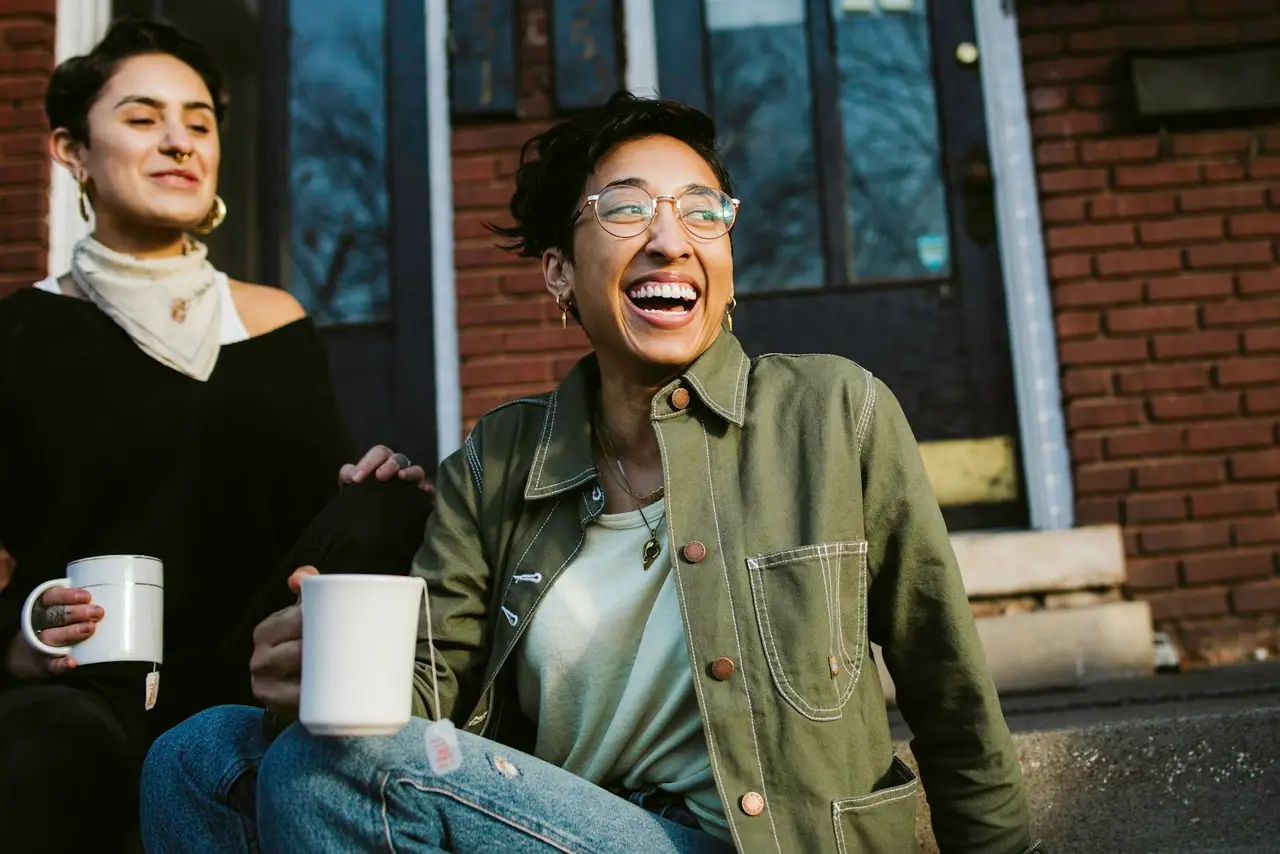 Two neighbors laughing over coffee on a front step.