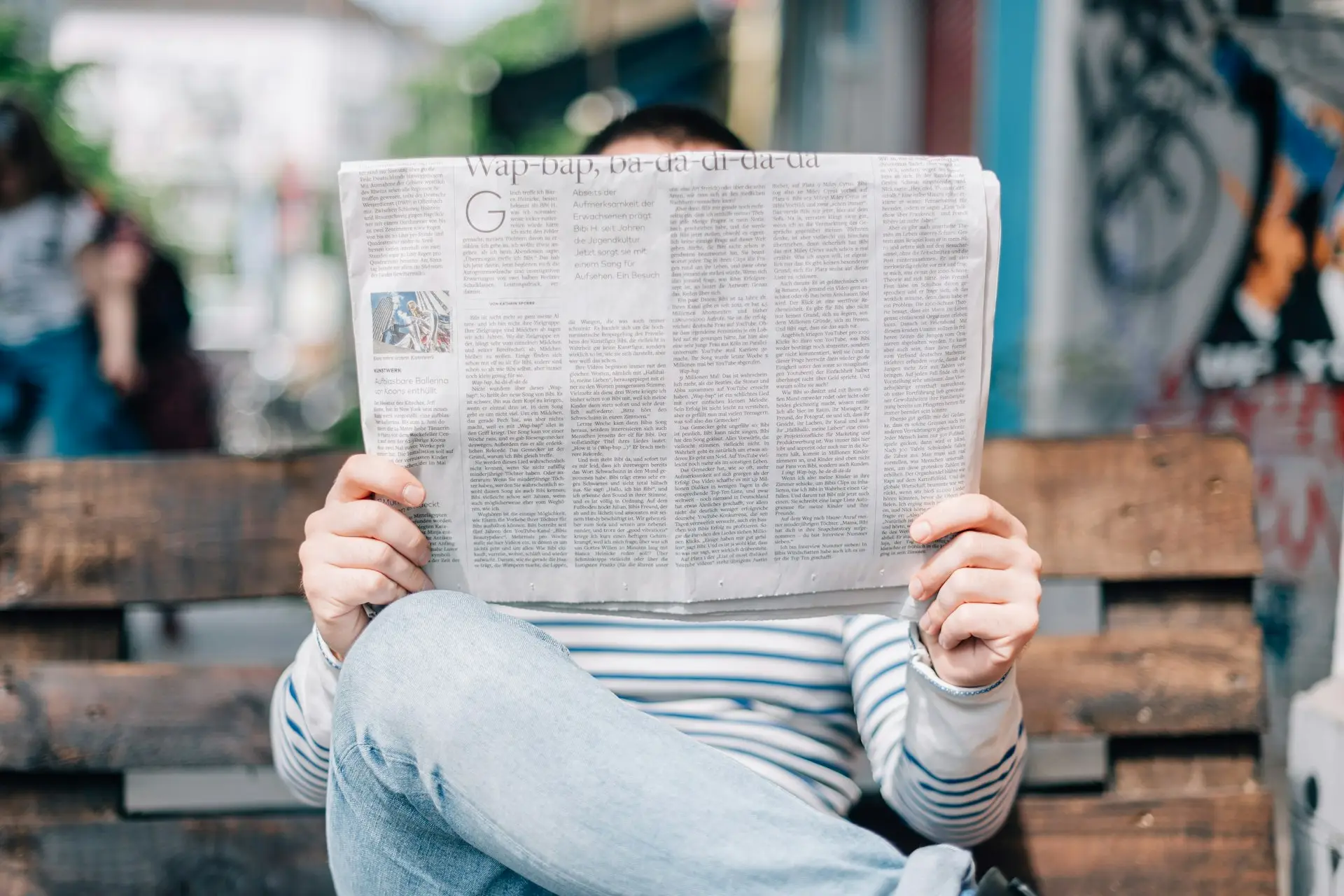 Eine Person sitzt auf einer Holzbank und liest eine aufgeschlagene Zeitung
