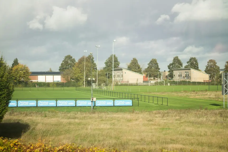 Rasensportplatz mit Flutlichtmasten, dahinter Wohn- und Schulgebäude
