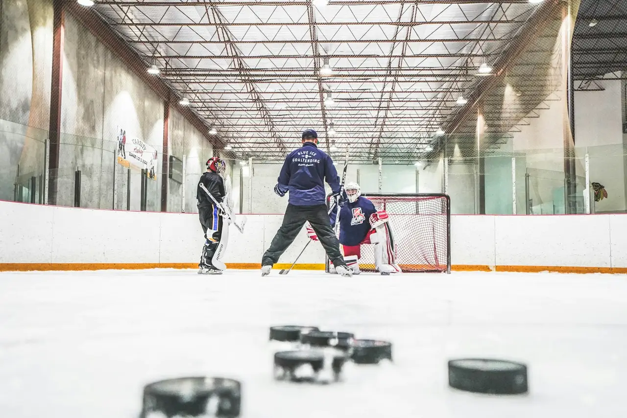 Ein Trainer steht beim Training mit einem Torwart am Tor, im Vordergrund liegen Pucks.