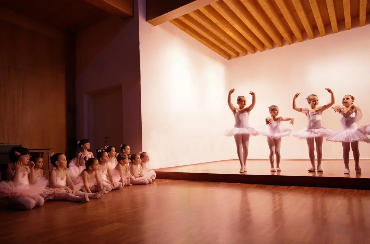 Kinder im Ballettkostüm tanzen in einem Saal, andere warten am Rand.