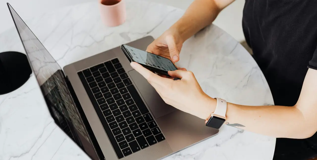 Frau sitzt vor einem Laptop und hält ein Handy in der Hand.