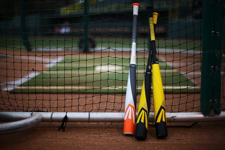 Drei Baseball-Schläger angelehnt ans Netz mit einem Baseballfeld im Hintergrund