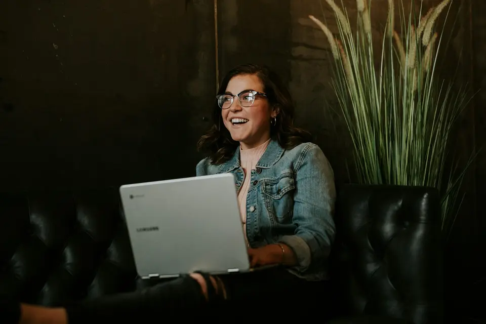 Eine Frau arbeitet lächelnd auf einem Ledersofa am Laptop.