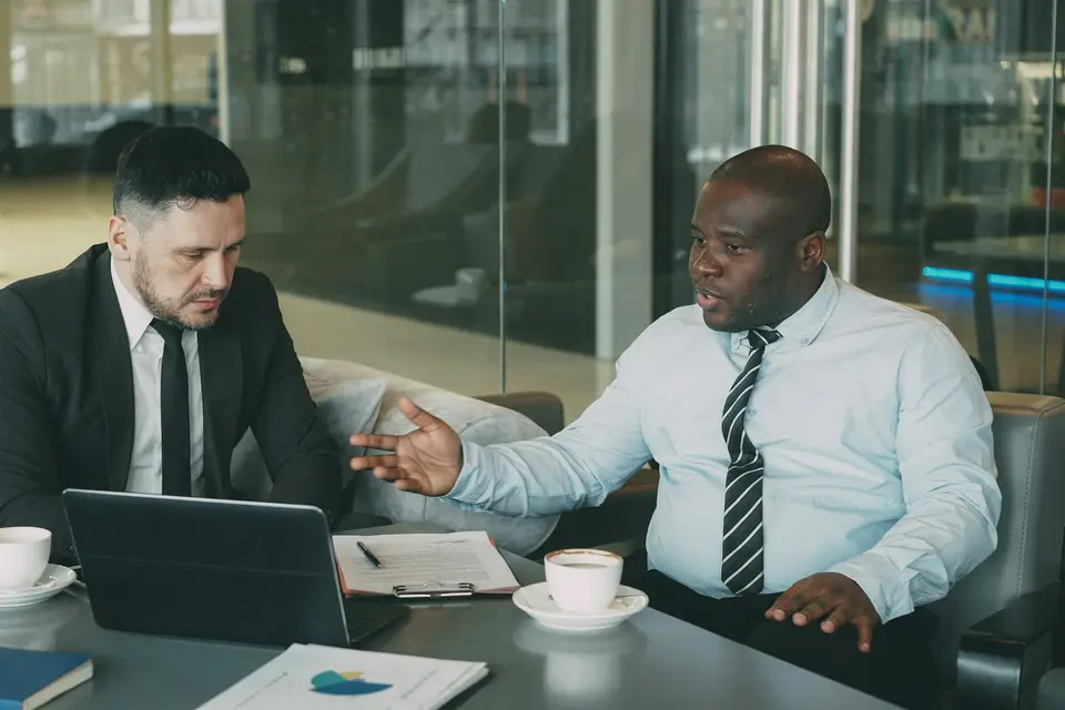 Zwei Männer besprechen an einem Tisch mit Laptop und Unterlagen eine Vereinbarung